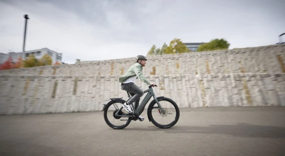 Ein Mann mit Helm fährt auf einem E-Bike vor einer Steinmauer, während der Himmel wolkenverhangen ist.