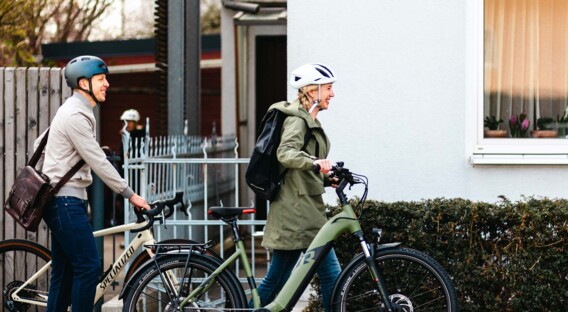 Ein Mann und eine Frau, beide mit Fahrradhelmen, schieben ihre Fahrräder entlang eines Gehwegs. Die Frau trägt einen Rucksack, und beide scheinen auf dem Weg zu sein.