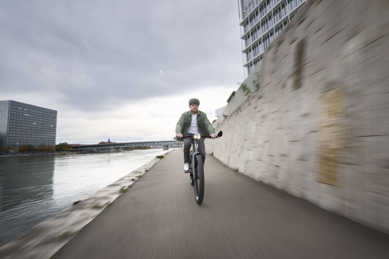 Ein Mann mit Helm fährt auf einem E-Bike entlang eines Flusses, vorbei an einer hohen Mauer, während der Himmel bewölkt ist.