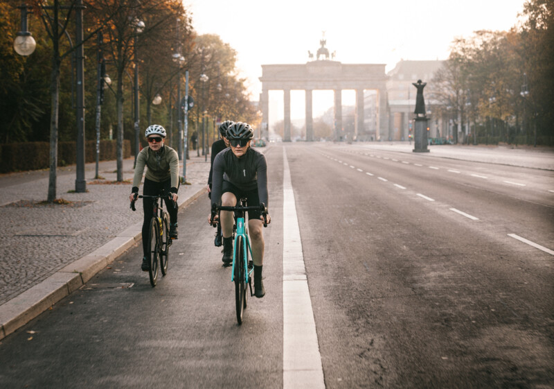 Drei Rennradfahrende fahren auf einer breiten, fast leeren Straße in Richtung Brandenburger Tor im Morgenlicht.