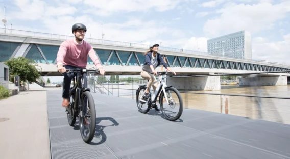 Eine Frau und ein Mann, beide mit Fahrradhelmen, unterhalten sich lächelnd. Die Frau steht neben einem E-Bike, während der Mann auf einer Bank sitzt, neben ihm ein Rennrad.