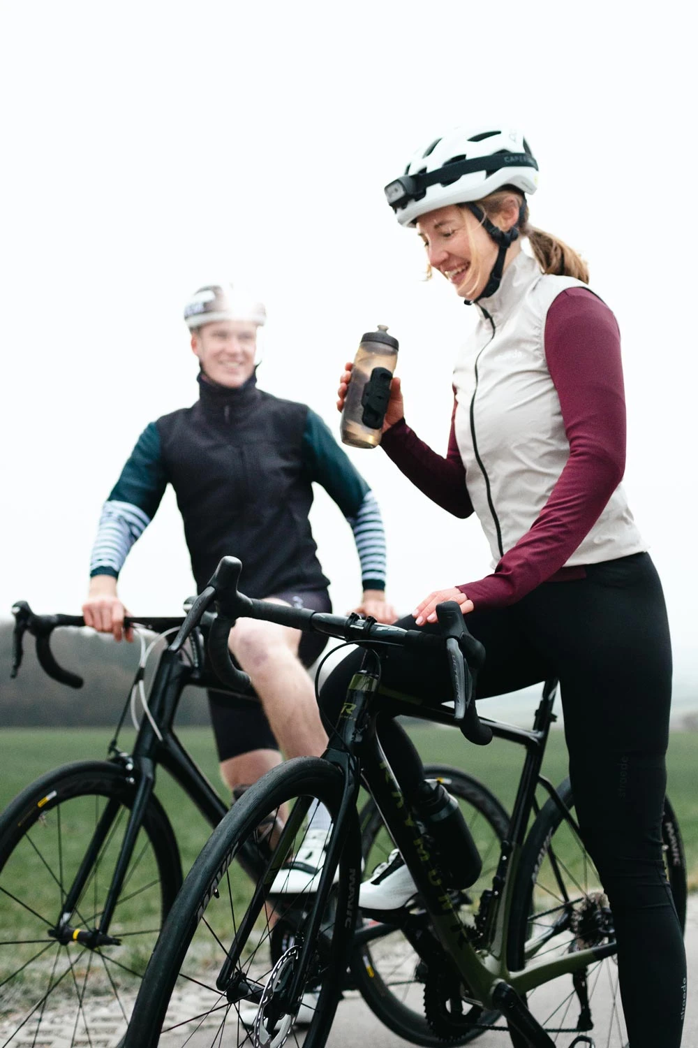 Zwei Radfahrer in Sportkleidung und Helmen machen eine Pause auf ihren Rennrädern. Eine Frau trinkt aus einer Wasserflasche, während der Mann im Hintergrund lächelnd sein Fahrrad hält.