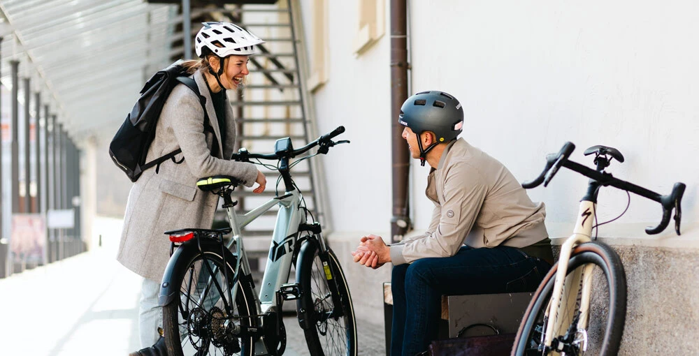 Eine Frau und ein Mann, beide mit Fahrradhelmen, unterhalten sich lächelnd. Die Frau steht neben einem E-Bike, während der Mann auf einer Bank sitzt, neben ihm ein Rennrad.