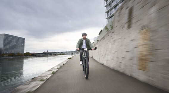 Ein Mann mit Helm fährt auf einem E-Bike entlang eines Flusses, vorbei an einer hohen Mauer, während der Himmel bewölkt ist.