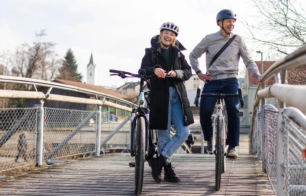 Ein Mann und eine Frau, beide mit Fahrradhelmen, stehen auf einer Brücke neben ihren Fahrrädern und lächeln. Im Hintergrund sind Bäume, Häuser und der Turm einer Kirche zu sehen.